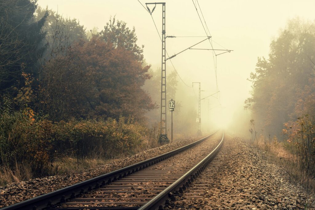 A foggy railway track stretches through a misty autumn landscape, lined with trees and gravel.